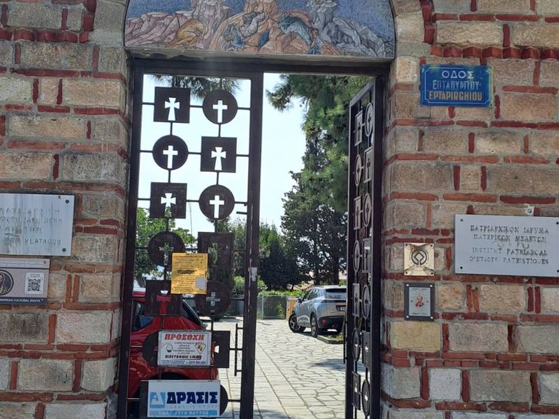 Πέτρινη τοξωτή πύλη με ψηφιδωτό της Μεταμόρφωσης πάνω από τη σιδερένια πόρτα με σταυρούς· μέσα διακρίνονται το πλακόστρωτο και δέντρα.
