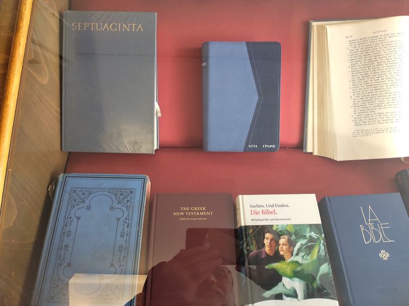 Γκρι τόμος «SEPTUAGINTA», μπλε Αγία Γραφή και ανοιχτή ελληνική σελίδα, πάνω από μικρότερες εκδόσεις σε κόκκινο φόντο.