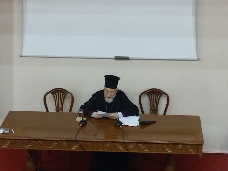 Κληρικός κοιτάζει τα χαρτιά που κρατά με τα δύο χέρια· πίσω του υπάρχουν δύο κενές ξύλινες καρέκλες.