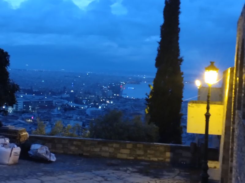 Ψηλό κυπαρίσσι στο κέντρο, αναμμένο φανάρι δεξιά και φωτισμένα κτίρια γύρω από τον κόλπο κάτω από συννεφιασμένο μπλε ουρανό.