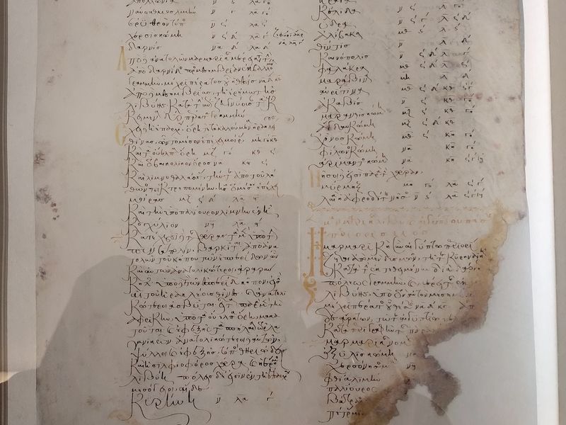 Αναπαραγωγή φύλλου με δύο στήλες ελληνικής γραφής, χρυσαφί αρχικά και μεγάλο φθαρμένο τμήμα στη δεξιά άκρη.