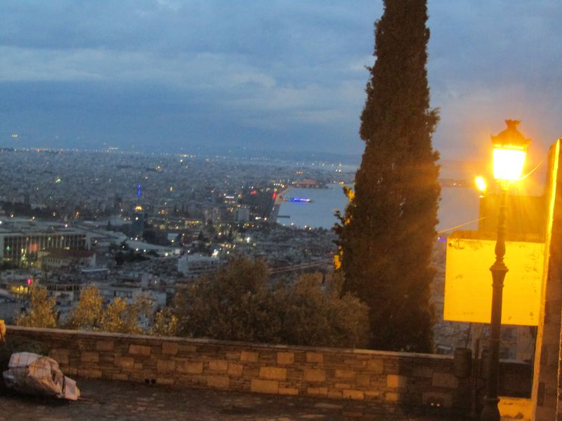 Φωτισμένο φανάρι και ψηλό κυπαρίσσι μπροστά σε πανοραμική θέα κτιρίων και θάλασσας.