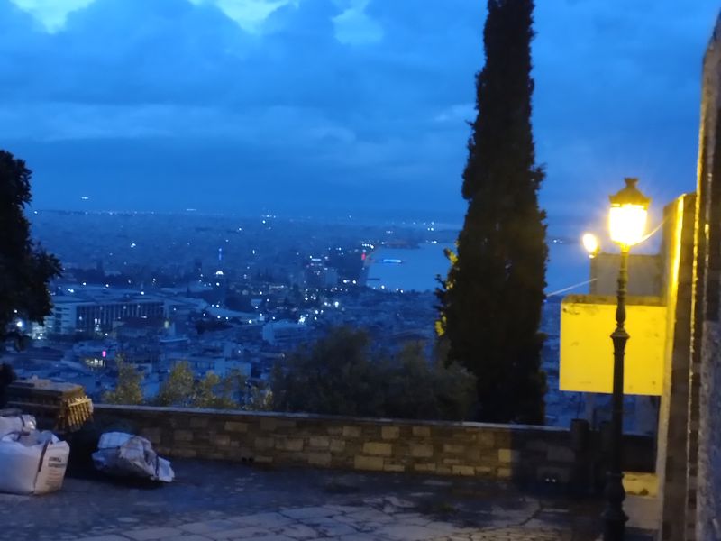 Κίτρινο φως λούζει την άκρη του πλακόστρωτου, ενώ ένα κυπαρίσσι προβάλλει μπροστά στην πόλη και στη θάλασσα.