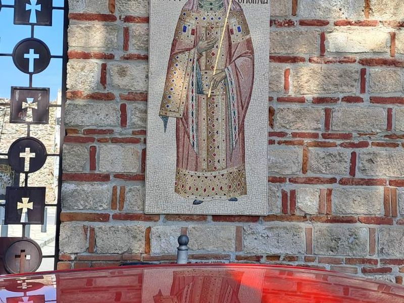 Η Άννα Παλαιολογίνα της Σαβοΐας με στέμμα, αυτοκρατορική ενδυμασία και σταυροφόρο σκήπτρο, κάτω από κυκλικό ψηφιδωτό με λευκό σταυρό σε κόκκινο φόντο· η μορφή αντανακλάται στην οροφή κόκκινου αυτοκινήτου.