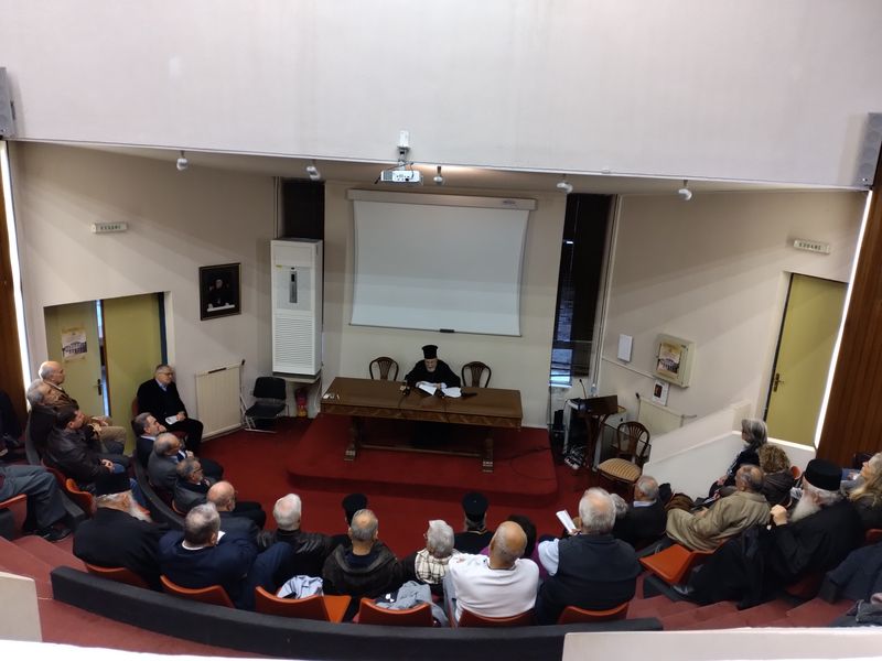 Άποψη από ψηλά: άνθρωποι σε καμπύλες σειρές καθισμάτων κοιτούν τον ομιλητή, που κάθεται κάτω από λευκή οθόνη.