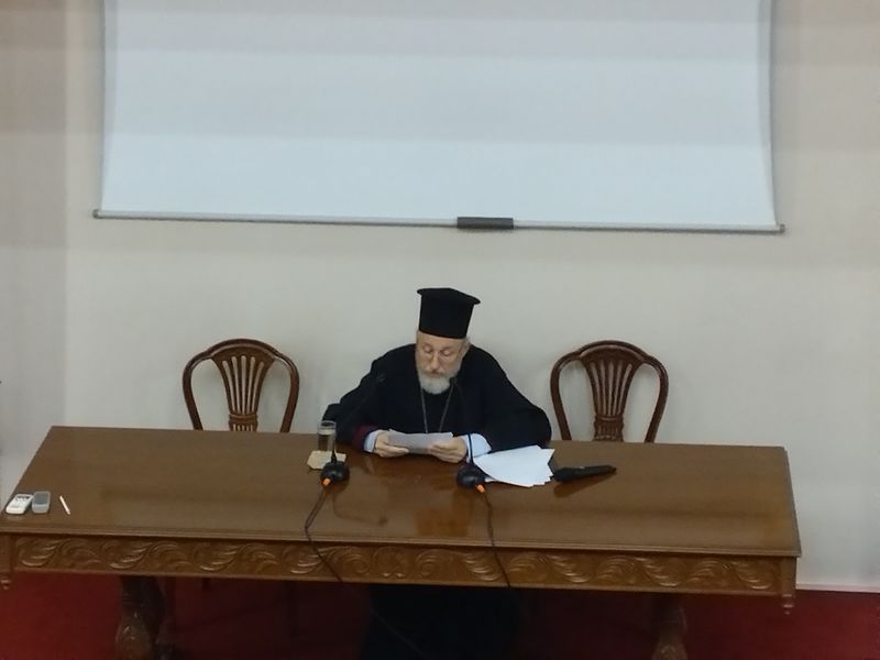 Κληρικός με γυαλιά κρατά ένα φύλλο πάνω από σκαλιστό τραπέζι, με μικρόφωνα και πρόσθετες σελίδες δίπλα του.
