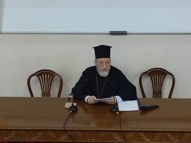 Κληρικός με μαύρο κάλυμμα κεφαλής και λευκή γενειάδα κοιτάζει μπροστά, κρατώντας χαρτιά ανάμεσα σε δύο μικρόφωνα.