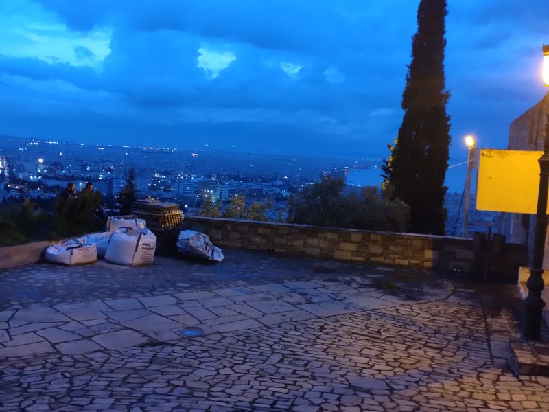 Πλακόστρωτη αυλή με λευκούς σάκους και κυπαρίσσι, με τα φώτα της πόλης και τον κόλπο στο βάθος.