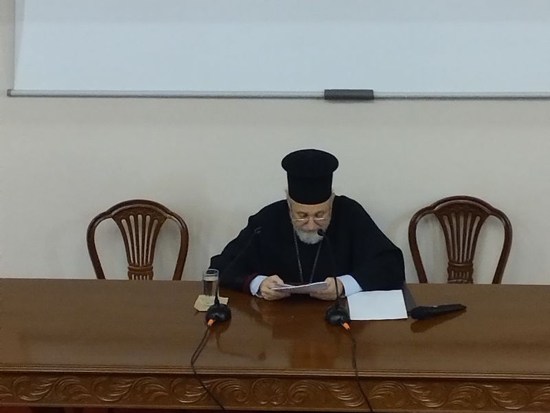 Καθισμένος κληρικός σκύβει πάνω από τα χαρτιά του, με δύο μικρόφωνα και ένα ποτήρι νερό στο ξύλινο τραπέζι.
