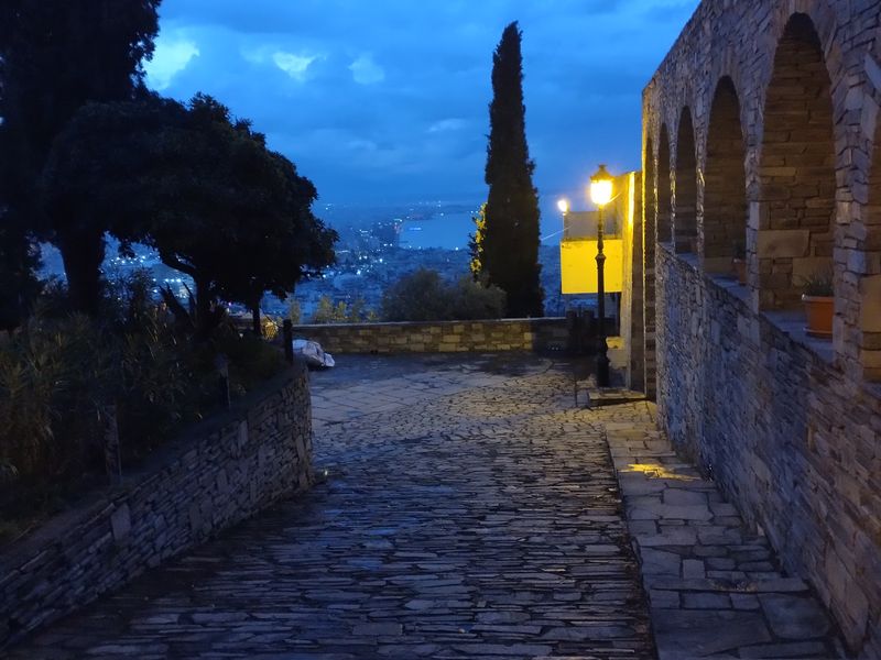 Πλακόστρωτο κατηφορίζει ανάμεσα σε δέντρα και πέτρινη στοά, προς αναμμένο φανάρι με την πόλη και τη θάλασσα πίσω του.