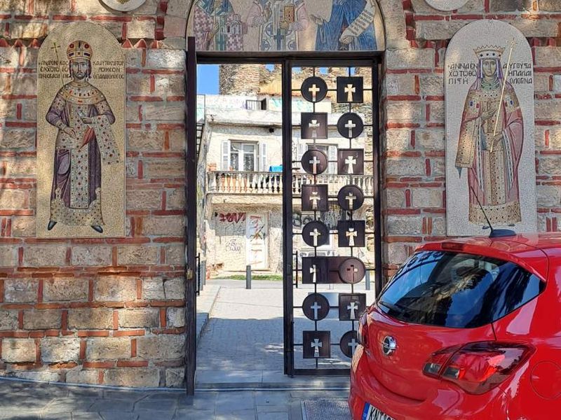 Πύλη από πέτρα και κόκκινα τούβλα, με σταυρό στην κορυφή, ψηφιδωτό τριών μορφών πάνω από το άνοιγμα και δύο αυτοκρατορικές μορφές στα πλάγια· κόκκινο αυτοκίνητο δεξιά.